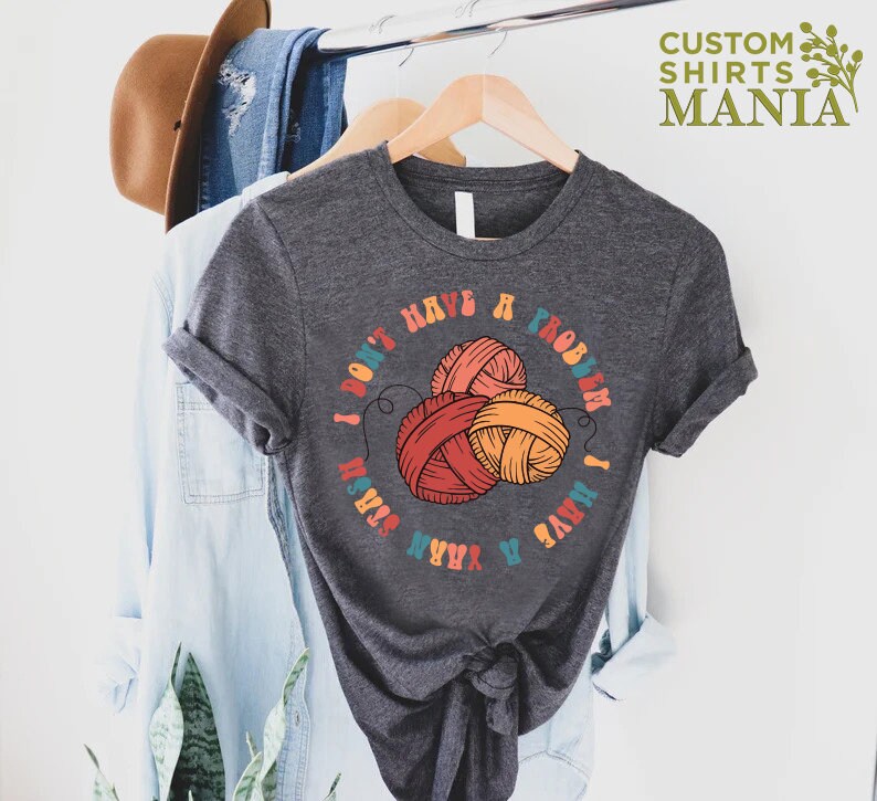 May include: A dark gray t-shirt with a colorful graphic that says "I don't have a problem I have yarn" with a circle of yarn balls in the center.