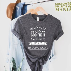 May include: A dark gray t-shirt with white text that reads "I'm going to let... GOD FIX IT because if I FIX IT I'M GOING TO JAIL".