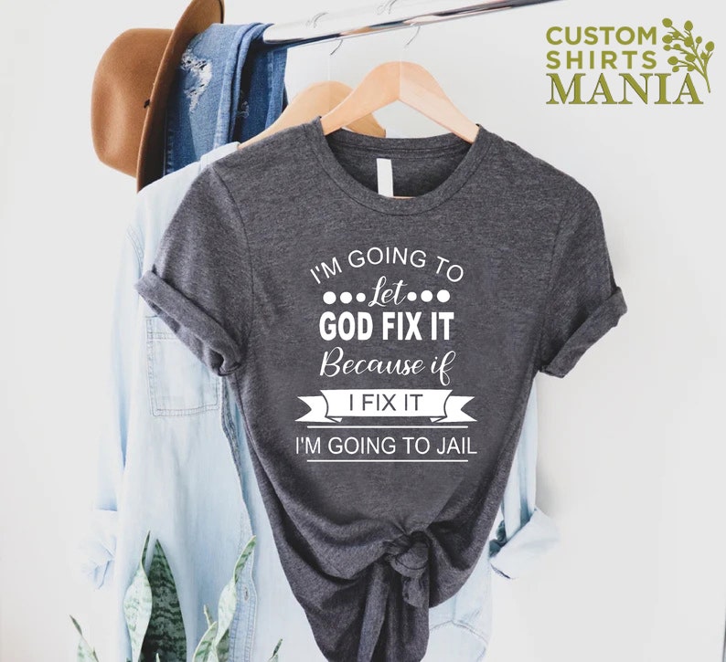 May include: A dark gray t-shirt with white text that reads "I'm going to let... GOD FIX IT because if I FIX IT I'M GOING TO JAIL".