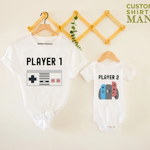 May include: Two white t-shirts with a video game theme. The larger shirt says "PLAYER 1" with a retro video game controller graphic. The smaller shirt says "PLAYER 2" with a Nintendo Switch controller graphic.