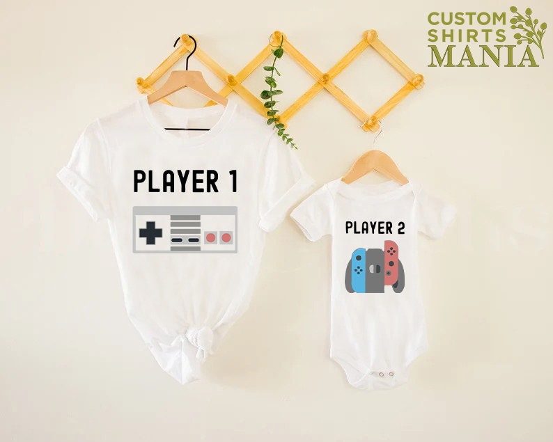 May include: Two white t-shirts with a video game theme. The larger shirt says "PLAYER 1" with a retro video game controller graphic. The smaller shirt says "PLAYER 2" with a Nintendo Switch controller graphic.