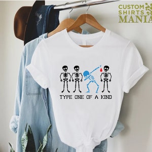 以下が含まれることがあります： 白いTシャツに、4つの骸骨のグラフィックがプリントされています。1つの骸骨はダブをしていて、頭が青く、赤い血の滴が落ちています。骸骨の下には「Type One Of A Kind」というテキストが書かれています。