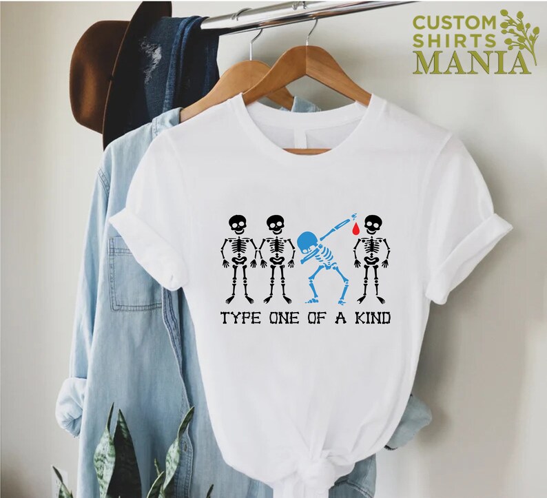 以下が含まれることがあります： 白いTシャツに、4つの骸骨のグラフィックがプリントされています。1つの骸骨はダブをしていて、頭が青く、赤い血の滴が落ちています。骸骨の下には「Type One Of A Kind」というテキストが書かれています。