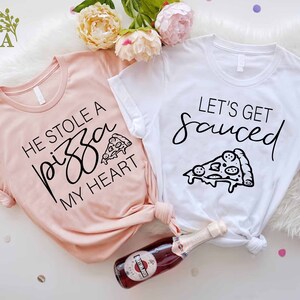Puede incluir: Dos camisetas con tem&aacute;tica de pizza. Una camiseta es rosa y dice "Se rob&oacute; una pizza de mi coraz&oacute;n" con un gr&aacute;fico de una rebanada de pizza. La otra camiseta es blanca y dice "Vamos a emborracharnos" con un gr&aacute;fico de una rebanada de pizza.