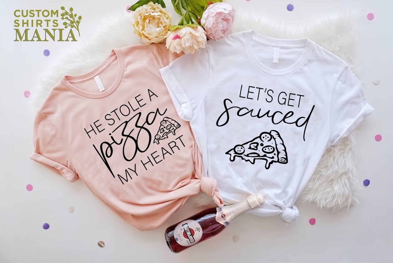 Puede incluir: Dos camisetas con tem&aacute;tica de pizza. Una camiseta es rosa y dice "Se rob&oacute; una pizza de mi coraz&oacute;n" con un gr&aacute;fico de una rebanada de pizza. La otra camiseta es blanca y dice "Vamos a emborracharnos" con un gr&aacute;fico de una rebanada de pizza.