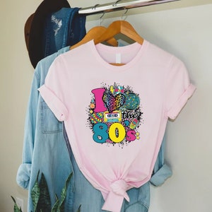 May include: Pink t-shirt with a graphic design of a heart with a leopard print, a disco ball, a cassette tape, a Rubik's cube, and the text "I love the 80's".