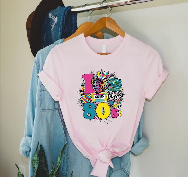 May include: Pink t-shirt with a graphic design of a heart with a leopard print, a disco ball, a cassette tape, a Rubik's cube, and the text "I love the 80's".