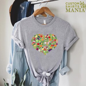 May include: A heather gray t-shirt featuring a heart-shaped design filled with colorful illustrations of various vegetables. The shirt has short sleeves and is hanging on a wooden hanger. The text "CUSTOM SHIRTS MANIA" is visible in the upper right corner.