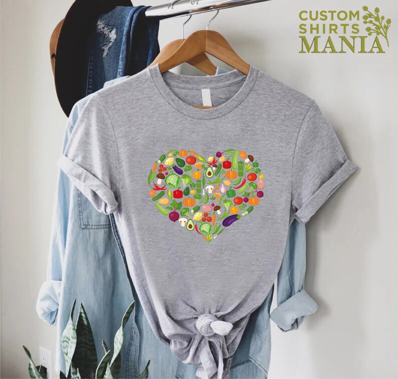 May include: A heather gray t-shirt featuring a heart-shaped design filled with colorful illustrations of various vegetables. The shirt has short sleeves and is hanging on a wooden hanger. The text "CUSTOM SHIRTS MANIA" is visible in the upper right corner.