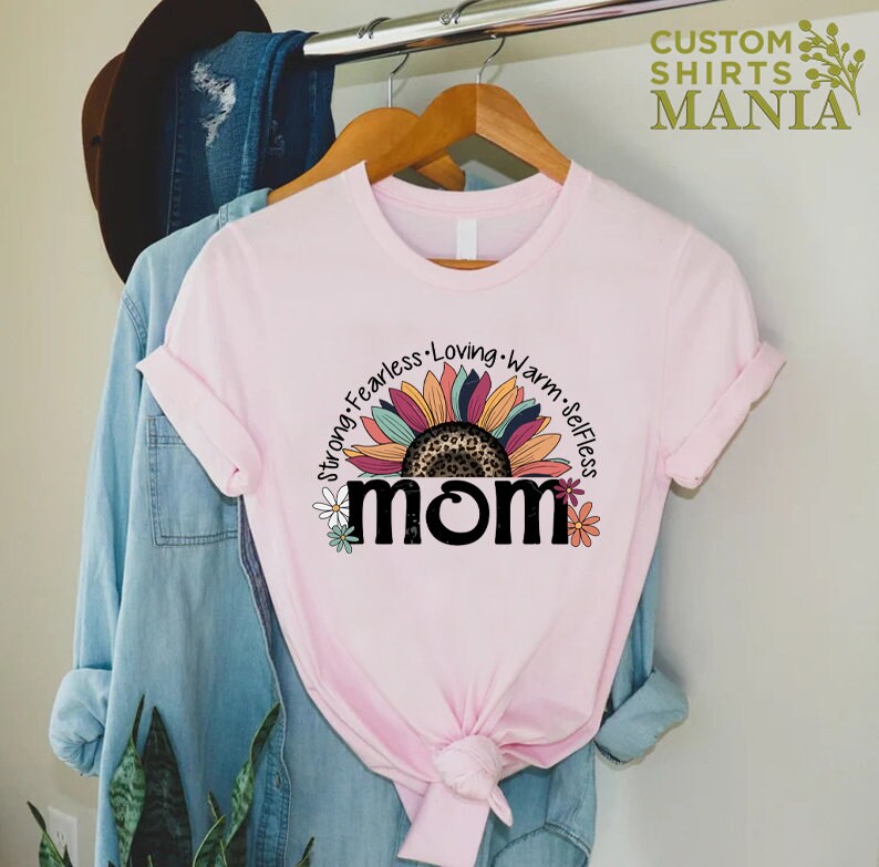 May include: Pink t-shirt featuring a sunflower graphic and the word "MOM" in black. The sunflower has a leopard print centre and the words "Strong, Fearless, Loving, Warm, Selfless" are written around the sunflower.