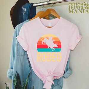 May include: Pink t-shirt with a vintage-style graphic of a cowboy riding a bull in front of a sunset. The text "RODEO" is printed below the graphic.