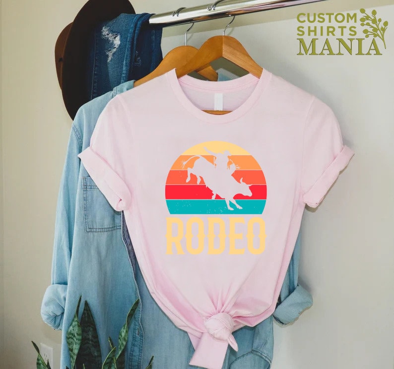 May include: Pink t-shirt with a vintage-style graphic of a cowboy riding a bull in front of a sunset. The text "RODEO" is printed below the graphic.