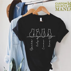 May include: Black t-shirt with a white outline of four cats sitting in a row. The names "Sparkle", "Lily", "Cosmo", and "Luna" are written underneath the cats.