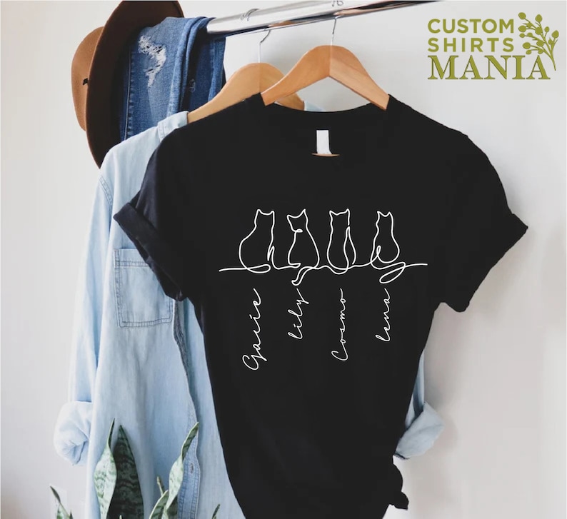 May include: Black t-shirt with a white outline of four cats sitting in a row. The names "Sparkle", "Lily", "Cosmo", and "Luna" are written underneath the cats.