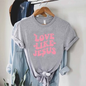 Może przedstawiać: Szary T-shirt z r&oacute;żowym nadrukiem, na kt&oacute;rym widnieje napis "LOVE LIKE JESUS".