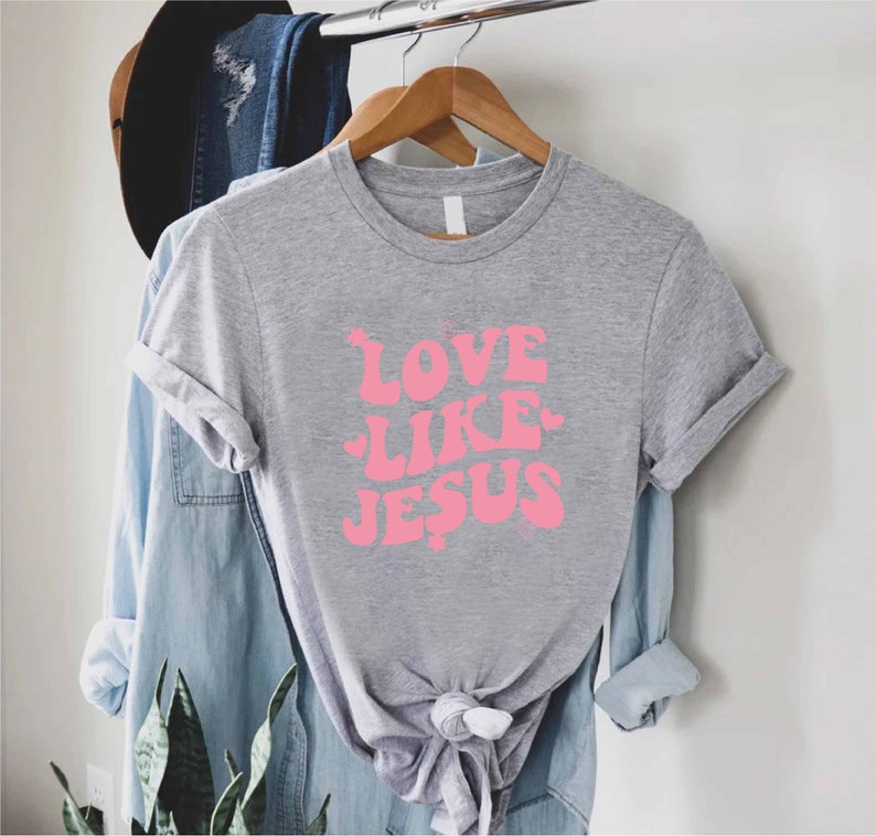 Może przedstawiać: Szary T-shirt z r&oacute;żowym nadrukiem, na kt&oacute;rym widnieje napis "LOVE LIKE JESUS".