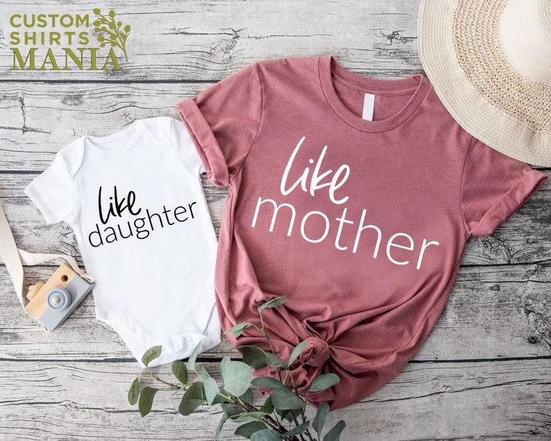 以下が含まれることがあります： 白とピンクの2枚のシャツのセットで、それぞれ「like daughter」と「like mother」の文字がプリントされています。シャツは木製の表面に置かれています。