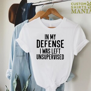May include: White t-shirt with black text that reads "In my defense I was left unsupervised".