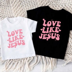 Może przedstawiać: Dwie koszulki z nadrukiem "Love Like Jesus" w r&oacute;żowym retro stylu na białym i czarnym tle.