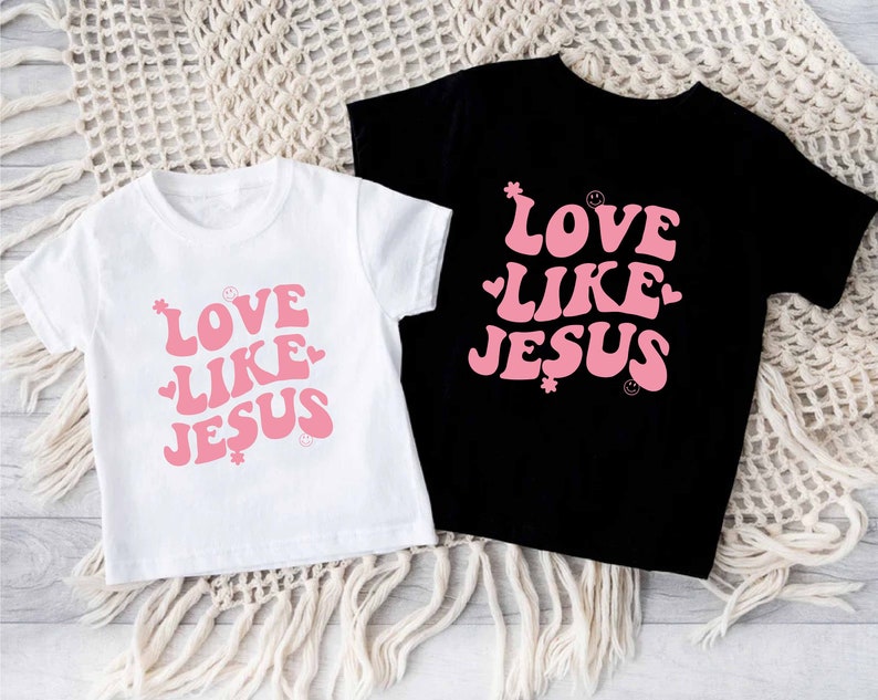 Może przedstawiać: Dwie koszulki z nadrukiem "Love Like Jesus" w r&oacute;żowym retro stylu na białym i czarnym tle.