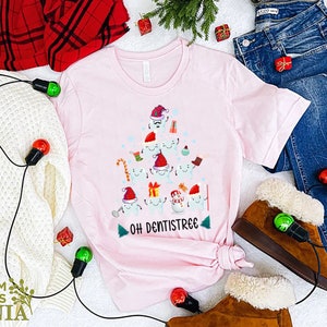以下が含まれることがあります： 歯でできたクリスマスツリーのデザインが施されたピンクのTシャツ。ツリーは、キャンディーケーン、プレゼント、オーナメントで飾られています。ツリーの下には「Oh Dentistree」という文字が印刷されています。