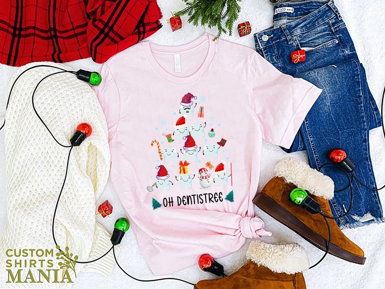 以下が含まれることがあります： 歯でできたクリスマスツリーのデザインが施されたピンクのTシャツ。ツリーは、キャンディーケーン、プレゼント、オーナメントで飾られています。ツリーの下には「Oh Dentistree」という文字が印刷されています。