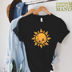 Puede incluir: Camiseta negra con un gr&aacute;fico de sol amarillo que presenta una mariposa y detalles florales. El sol tiene un aspecto estilizado y vintage.