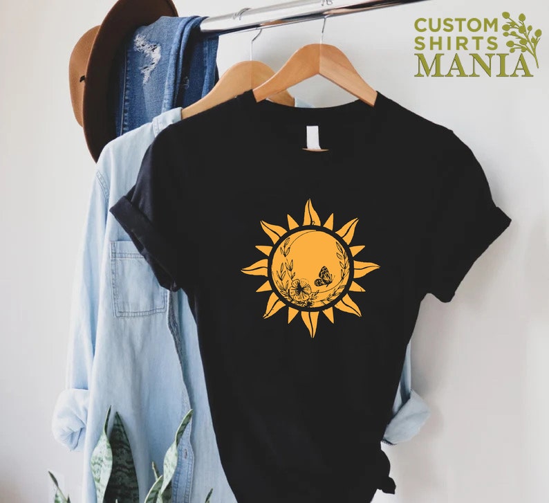 Puede incluir: Camiseta negra con un gr&aacute;fico de sol amarillo que presenta una mariposa y detalles florales. El sol tiene un aspecto estilizado y vintage.