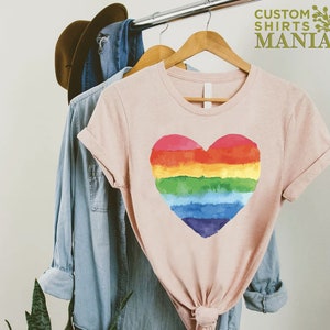 Peut inclure: T-shirt rose clair avec un motif de c&oelig;ur arc-en-ciel peint &agrave; l'aquarelle. Le t-shirt est suspendu &agrave; un cintre en bois avec d'autres v&ecirc;tements. Le texte "CUSTOM SHIRTS MANIA" est visible en haut &agrave; droite.