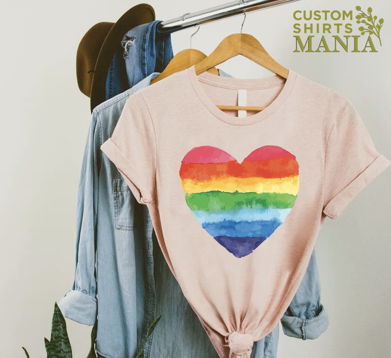 Peut inclure: T-shirt rose clair avec un motif de c&oelig;ur arc-en-ciel peint &agrave; l'aquarelle. Le t-shirt est suspendu &agrave; un cintre en bois avec d'autres v&ecirc;tements. Le texte "CUSTOM SHIRTS MANIA" est visible en haut &agrave; droite.