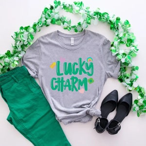May include: A grey t-shirt with the text "Lucky Charm" in green lettering. The shirt is decorated with small colourful shamrocks and a rainbow. The shirt is laid out on a white surface with green shamrock garland and green trousers.