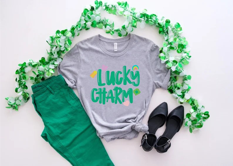 May include: A grey t-shirt with the text "Lucky Charm" in green lettering. The shirt is decorated with small colourful shamrocks and a rainbow. The shirt is laid out on a white surface with green shamrock garland and green trousers.