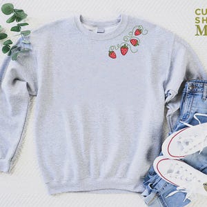 Puede incluir: Sudadera gris claro con fresas rojas bordadas y vides verdes. La sudadera se combina con vaqueros azules y zapatillas blancas. El texto "CUSTOM SHIRTS MANIA" es visible en la esquina superior derecha.