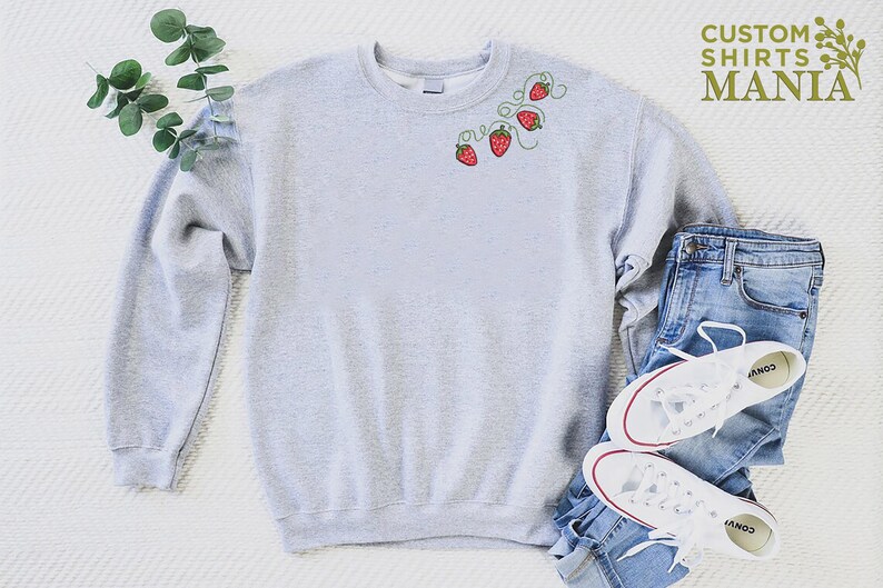 Puede incluir: Sudadera gris claro con fresas rojas bordadas y vides verdes. La sudadera se combina con vaqueros azules y zapatillas blancas. El texto "CUSTOM SHIRTS MANIA" es visible en la esquina superior derecha.