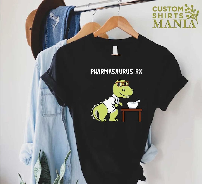 May include: Black t-shirt featuring a green dinosaur wearing glasses and a white lab coat. The dinosaur is holding a mortar and pestle. The text "PHARMASAURUS RX" is printed above the dinosaur.