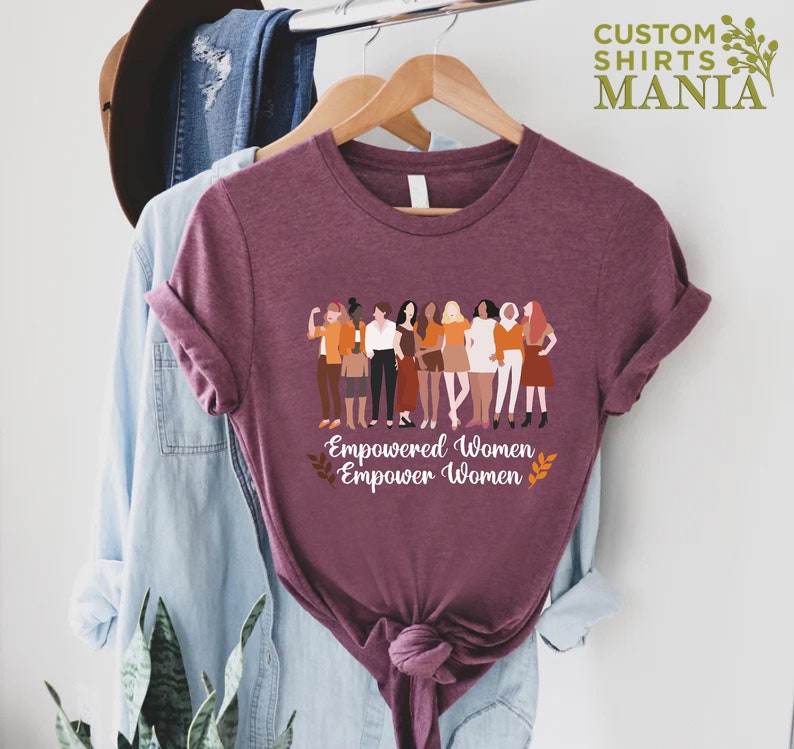 May include: A maroon t-shirt featuring a graphic of a group of diverse women standing together. The text "Empowered Women Empower Women" is printed below the graphic.