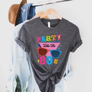 K&ouml;nnte beinhalten: Ein graues T-Shirt mit einem grafischen Design, das den Text "Party Like The 80s" in bunten Buchstaben und eine Sonnenbrille im Retro-Design zeigt.