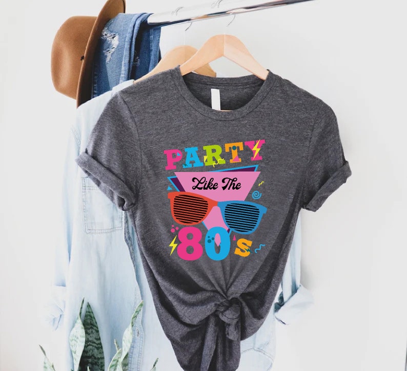 K&ouml;nnte beinhalten: Ein graues T-Shirt mit einem grafischen Design, das den Text "Party Like The 80s" in bunten Buchstaben und eine Sonnenbrille im Retro-Design zeigt.