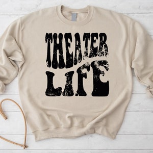 May include: A beige sweatshirt with the text "Theater Life" printed in black in a retro font.