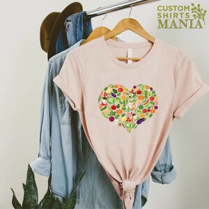 May include: A light pink t-shirt with a heart-shaped design made of various colorful vegetables. The shirt is hanging on a wooden hanger with other clothing items. The text "CUSTOM SHIRTS MANIA" is visible in the upper right corner.