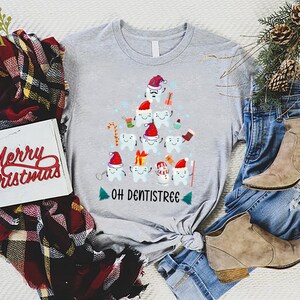 以下が含まれることがあります： サンタの帽子をかぶった歯でできたクリスマスツリーのデザインが施されたグレーのTシャツ。ツリーは、キャンディーケーン、プレゼント、歯ブラシで飾られています。ツリーの下には「Oh Dentistree」という文字があります。