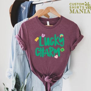 May include: A maroon t-shirt with the text "Lucky Charm" in green lettering. The shirt has a rainbow, a horseshoe, a four-leaf clover, and a heart graphic.