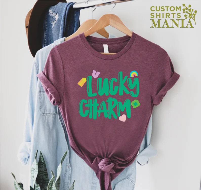 May include: A maroon t-shirt with the text "Lucky Charm" in green lettering. The shirt has a rainbow, a horseshoe, a four-leaf clover, and a heart graphic.