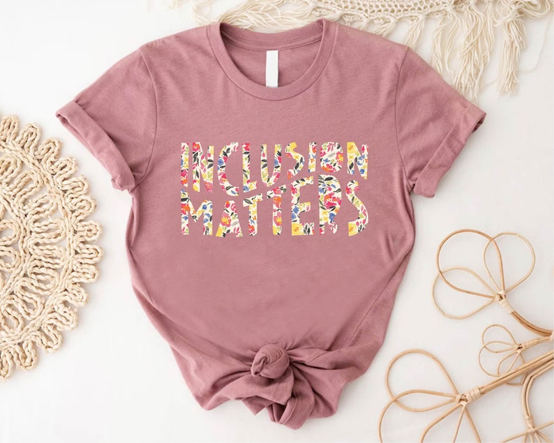 Puede incluir: Una camiseta de color malva con mangas cortas y un dise&ntilde;o de estampado floral que dice "Inclusion Matters".