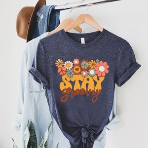 May include: A navy blue t-shirt with a retro-style graphic design featuring flowers, peace signs, and the text "Stay Groovy".