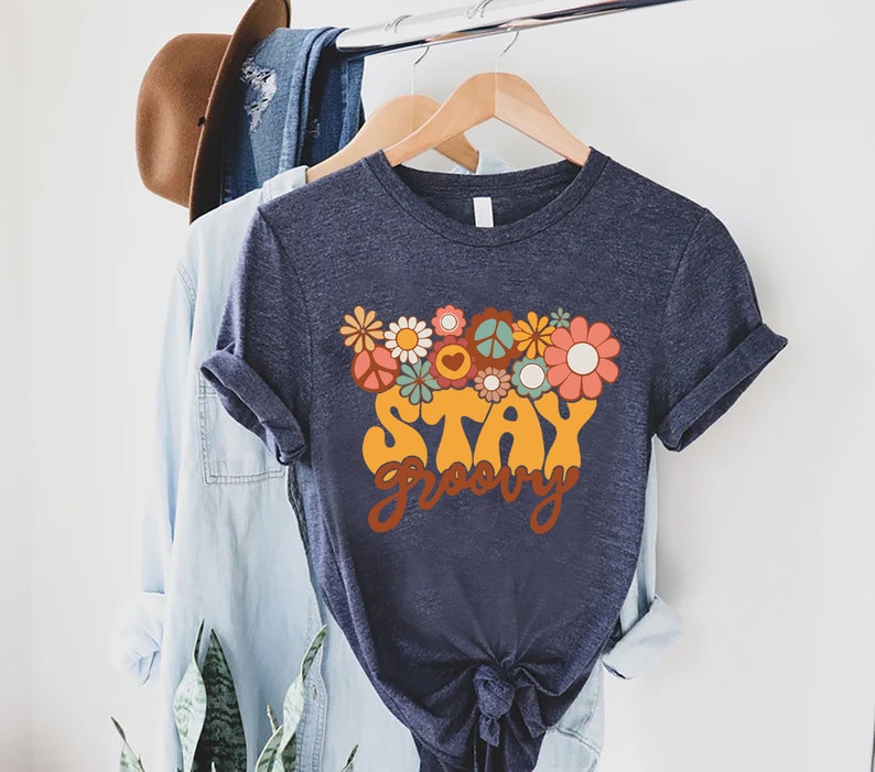 May include: A navy blue t-shirt with a retro-style graphic design featuring flowers, peace signs, and the text "Stay Groovy".