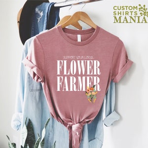 May include: A pink t-shirt with the text "Support Your Local Flower Farmer" in white lettering. The shirt has a floral design in the bottom right corner.