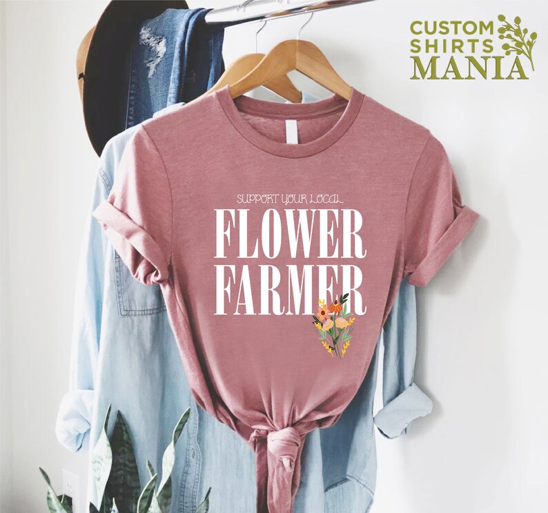 May include: A pink t-shirt with the text "Support Your Local Flower Farmer" in white lettering. The shirt has a floral design in the bottom right corner.