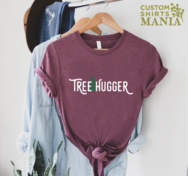 May include: A maroon t-shirt with the text "Tree Hugger" in white with a green tree graphic. The shirt is hanging on a wooden hanger.