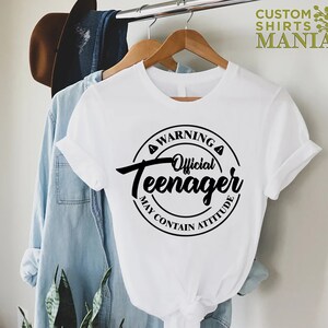 May include: White t-shirt with a black circle graphic that says "Warning Official Teenager May Contain Attitude".